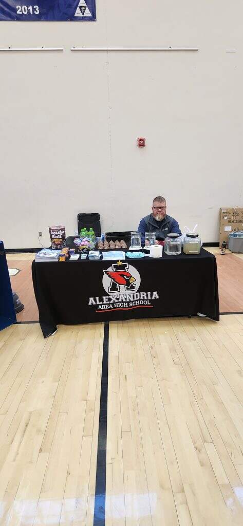 A person sits behind a table covered with an Alexandria Area High School cloth, displaying snacks, drinks, and informational items in a gymnasium setting.