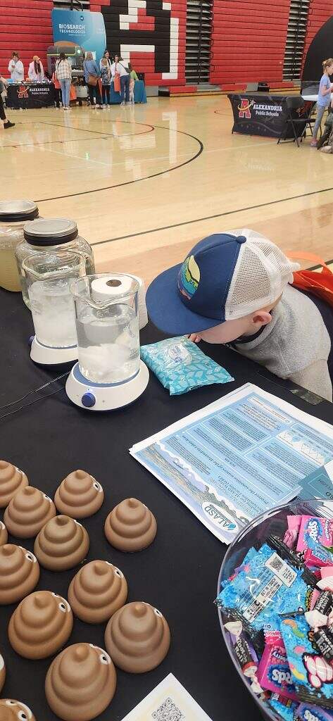 A young boy in a baseball cap leans over a table displaying fake poop, jars of water, a beaker on a hot plate, informational pamphlets, and candy at a science fair in a school gymnasium.