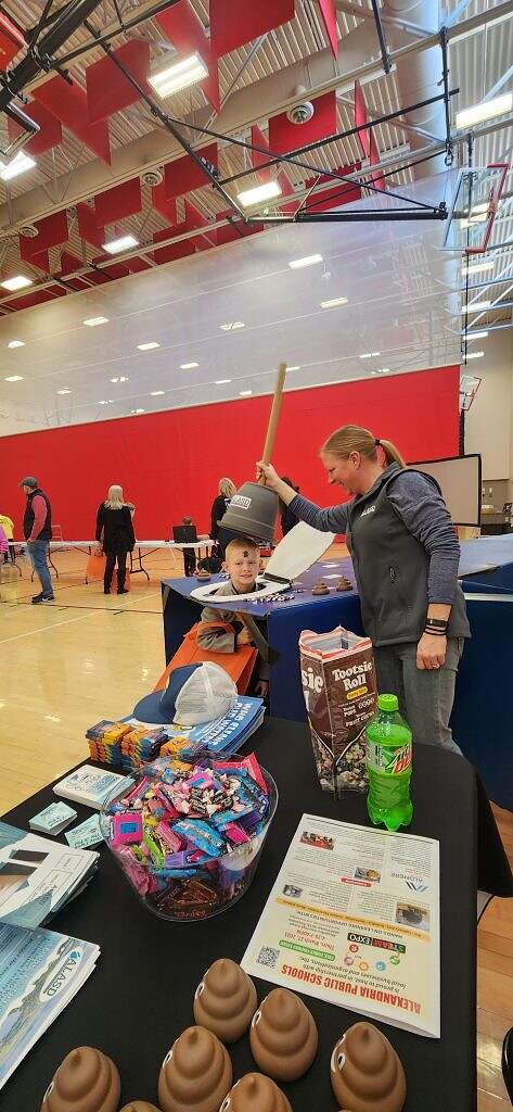 A woman playfully puts a toy trash can on a boy's head at a school gym event. The table in front of them holds candy, soda, brochures, and toy poop emojis. Other people are in the background.