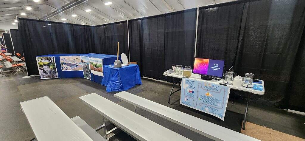 An indoor exhibit for groundwater education at the 2025 Kids Groundwater Festival features benches facing a display table with water jars, a monitor, and educational materials. Behind it, a blue table displays photos and a toilet model demonstrating water treatment.