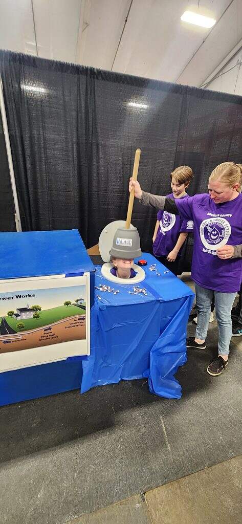 Two women in purple shirts stand beside a table with a blue cloth. One lowers a prop manhole cover onto someone's head poking through the table, demonstrating groundwater education at the 2025 Kids Groundwater Festival.