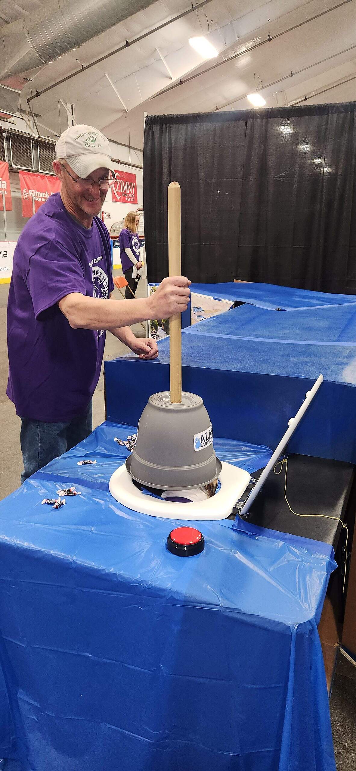A man wearing glasses and a purple shirt smiles while using a device with a long wooden handle and gray bucket at a booth covered with blue tablecloths during the 2025 Kids Groundwater Festival, promoting groundwater education in an indoor event space.