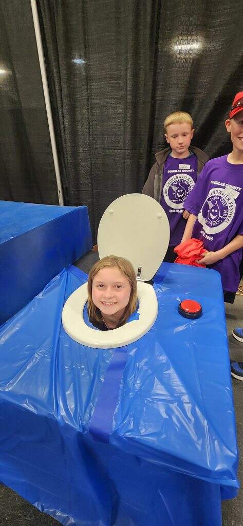 A smiling child’s head pokes through a hole in a table made to look like a toilet at the 2025 Groundwater Festival. Two other kids in purple shirts stand nearby, one holding a red object, all part of fun kids environmental education activities.