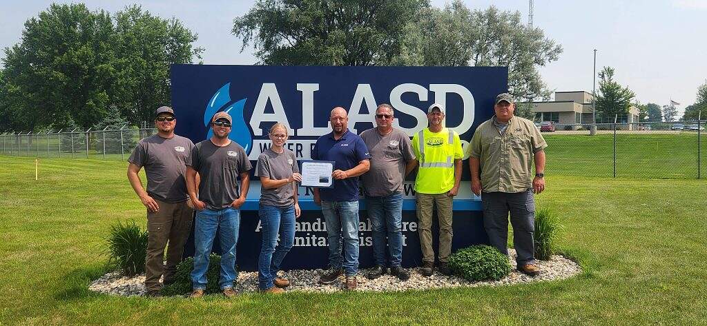 Seven people stand on grass in front of a large blue ALASD sign. One person in the center holds a certificate. Most are wearing work uniforms and safety gear. Trees and a building are visible in the background.