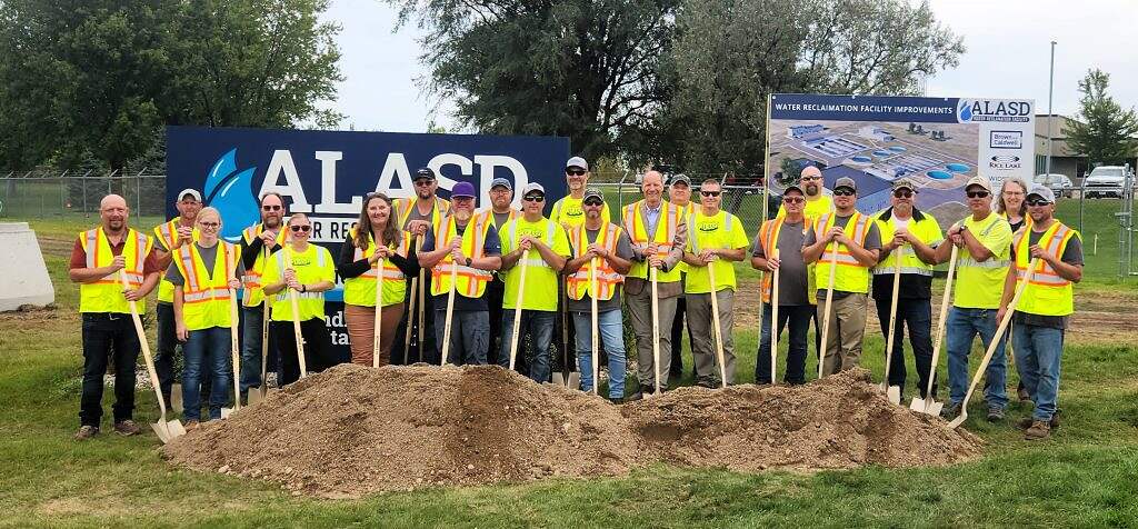 A group of people wearing yellow safety vests and helmets stand in a line, holding shovels at a groundbreaking ceremony for an $80 million infrastructure project. Behind them are signs for ALASD and a facility improvement project plan.