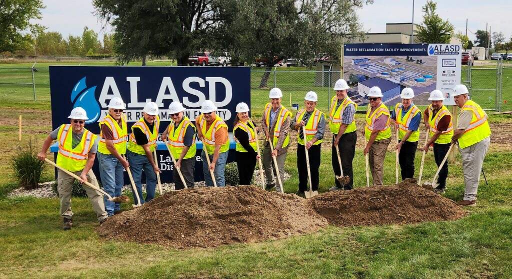 A group in safety vests and hard hats stands in a row, holding shovels at an $80 million infrastructure project groundbreaking. Behind them are ALASD signs and a facility improvement board, with dirt piles ready for the ceremony.