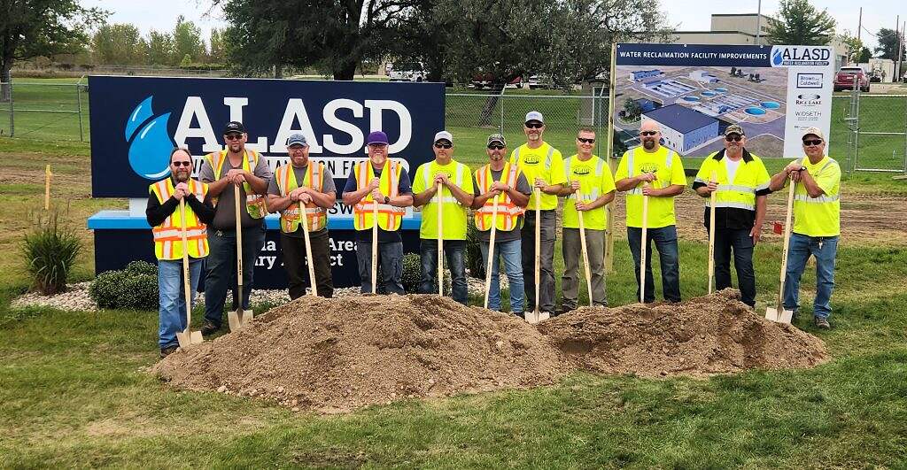 A group of people in yellow safety vests and hard hats stand in a row holding shovels at the $80 million infrastructure project groundbreaking ceremony, with a sign and facility rendering displayed behind them on a grassy area.