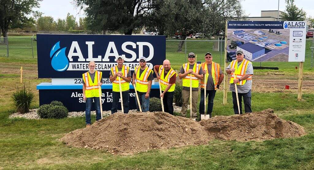 Eight people in safety vests stand with shovels at a groundbreaking event for the $80 million ALASD Water Reclamation Facility, with a project display behind them and dirt piles and grass in the foreground.
