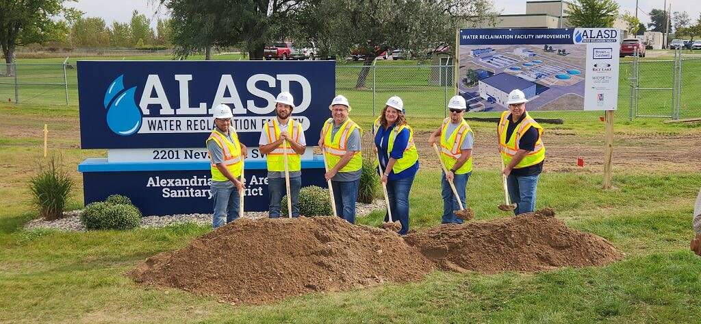 Six people in safety vests and hard hats pose with shovels for a groundbreaking ceremony, celebrating an $80 million infrastructure project at the ALASD Water Reclamation Facility, with a project rendering, grass, trees, and fence in the background.