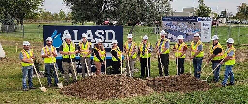 A group of people wearing hard hats and safety vests stand in a line holding shovels at the $80 million infrastructure project groundbreaking, with an ALASD sign and construction plans in the background.