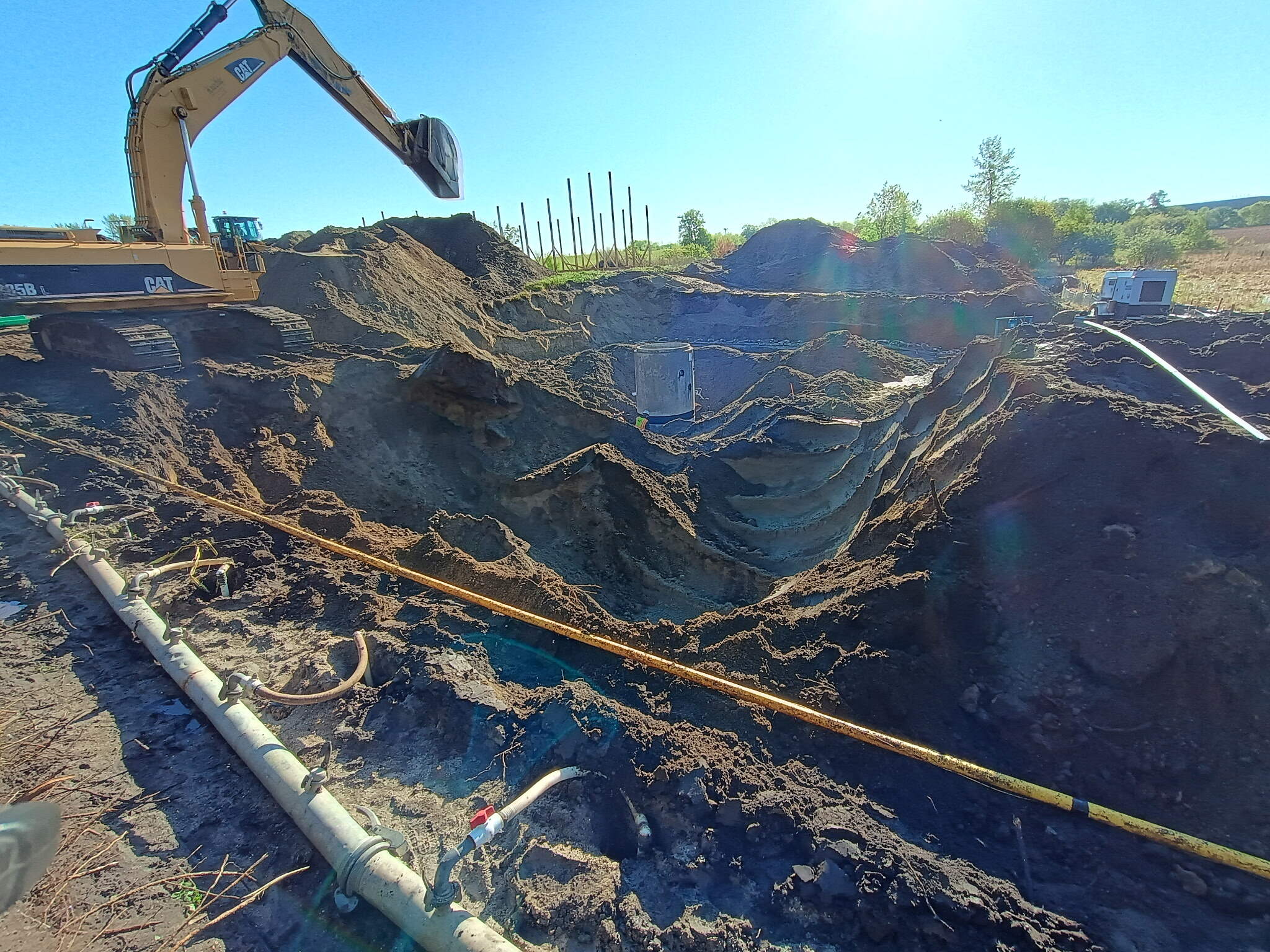 A large construction excavator sits by a deep, stepped excavation pit at the ALASR Extension Project. Pipes and cables run along the ground, with utility boxes visible inside the pit. The scene is outdoors on a sunny day.