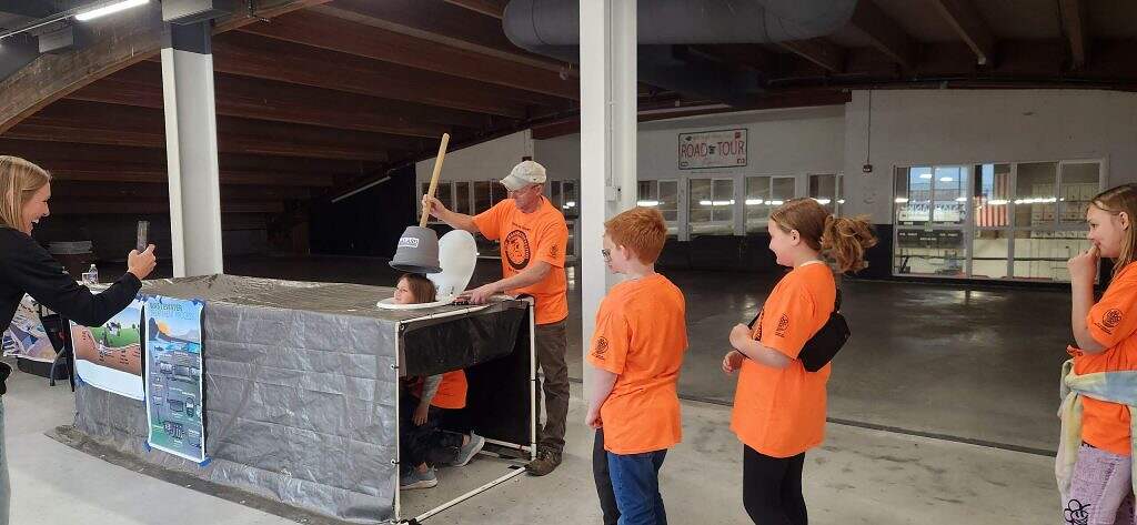 A group of kids in orange shirts watch as one child, wearing a helmet, sits with their head through a table hole while another person prepares to lift a toilet seat over their head at the Ground Water Festival. A woman takes a photo with her phone.