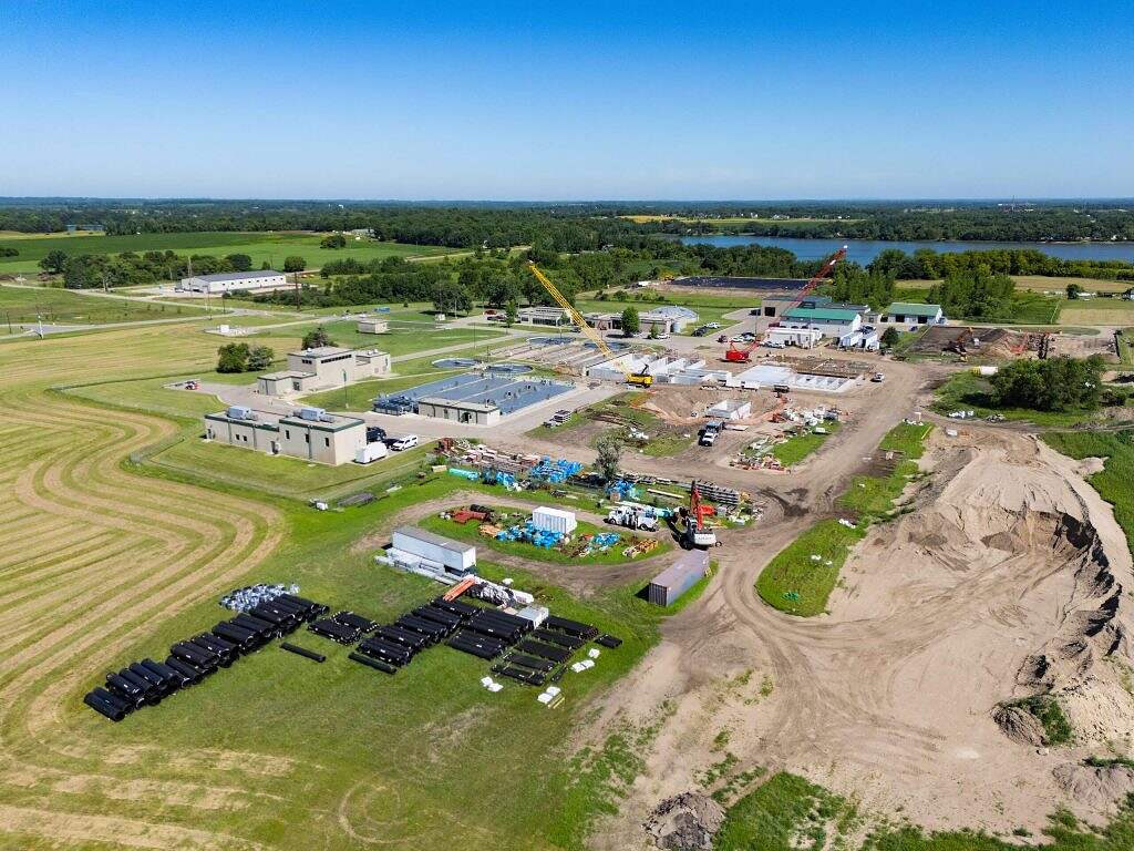 Aerial view of a construction site showcasing ongoing Construction and WRF Improvements, with cranes, buildings, equipment, and materials arranged on open land, surrounded by grassy fields and distant trees under a clear blue sky.