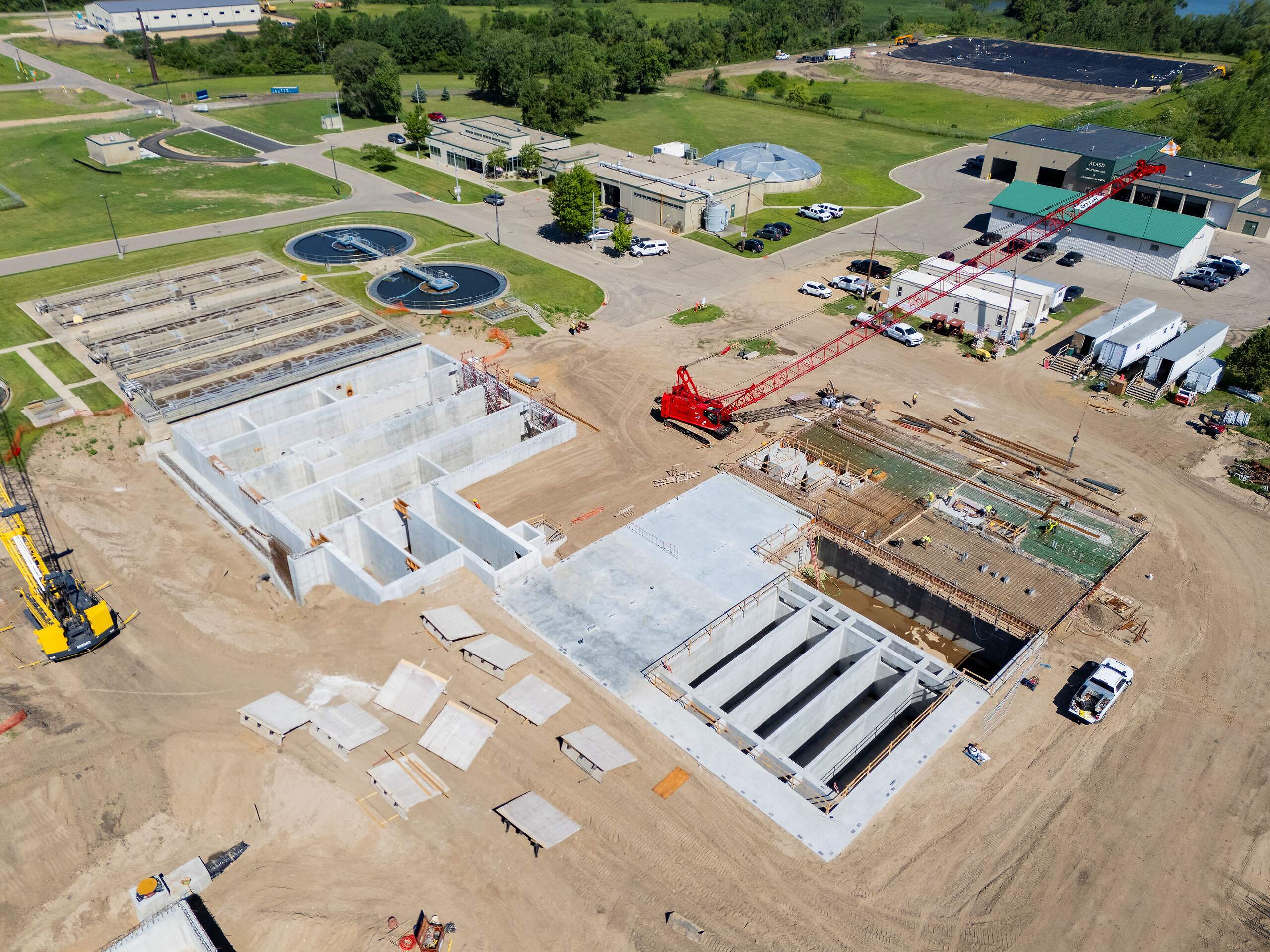 Aerial view of a construction site for Water Reclamation Facility (WRF) Improvements, featuring large concrete structures, cranes, vehicles, and nearby completed buildings surrounded by grass and roads.