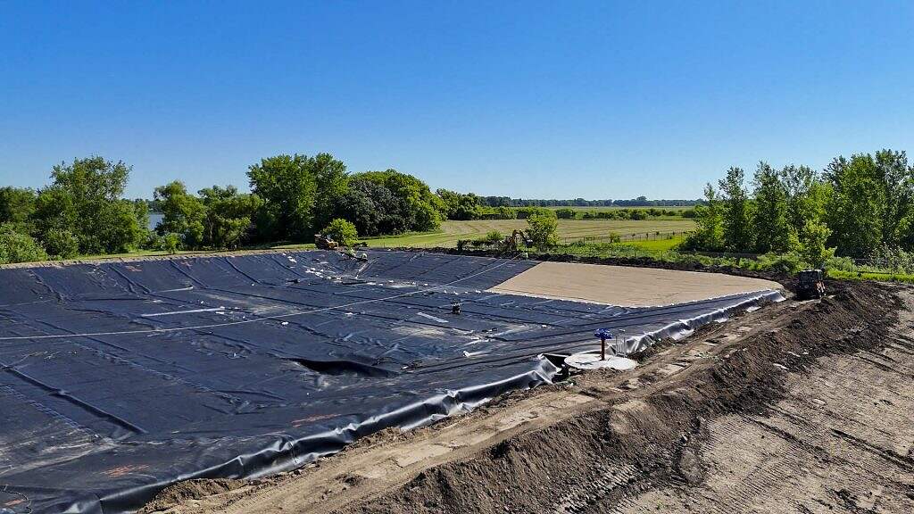 A large outdoor equalization basin, often used as part of a WRF (Water Reclamation Facility), is lined with black plastic tarp and sits in a rural area surrounded by trees and open fields under a clear blue sky.