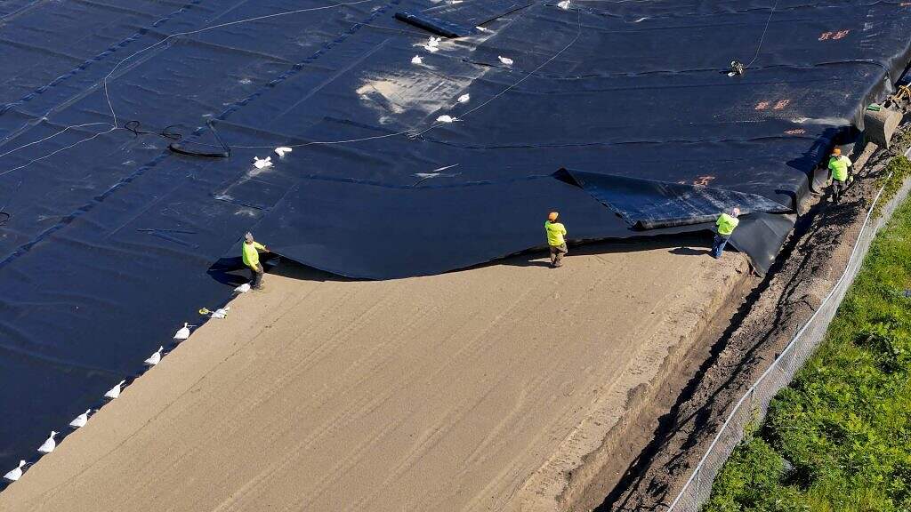 Three workers in safety gear install large black plastic sheeting over a sandy surface at a construction site, bordering an area with green vegetation, as part of preparations for constructing an Equalization Basin at the Water Resource Facility (WRF).