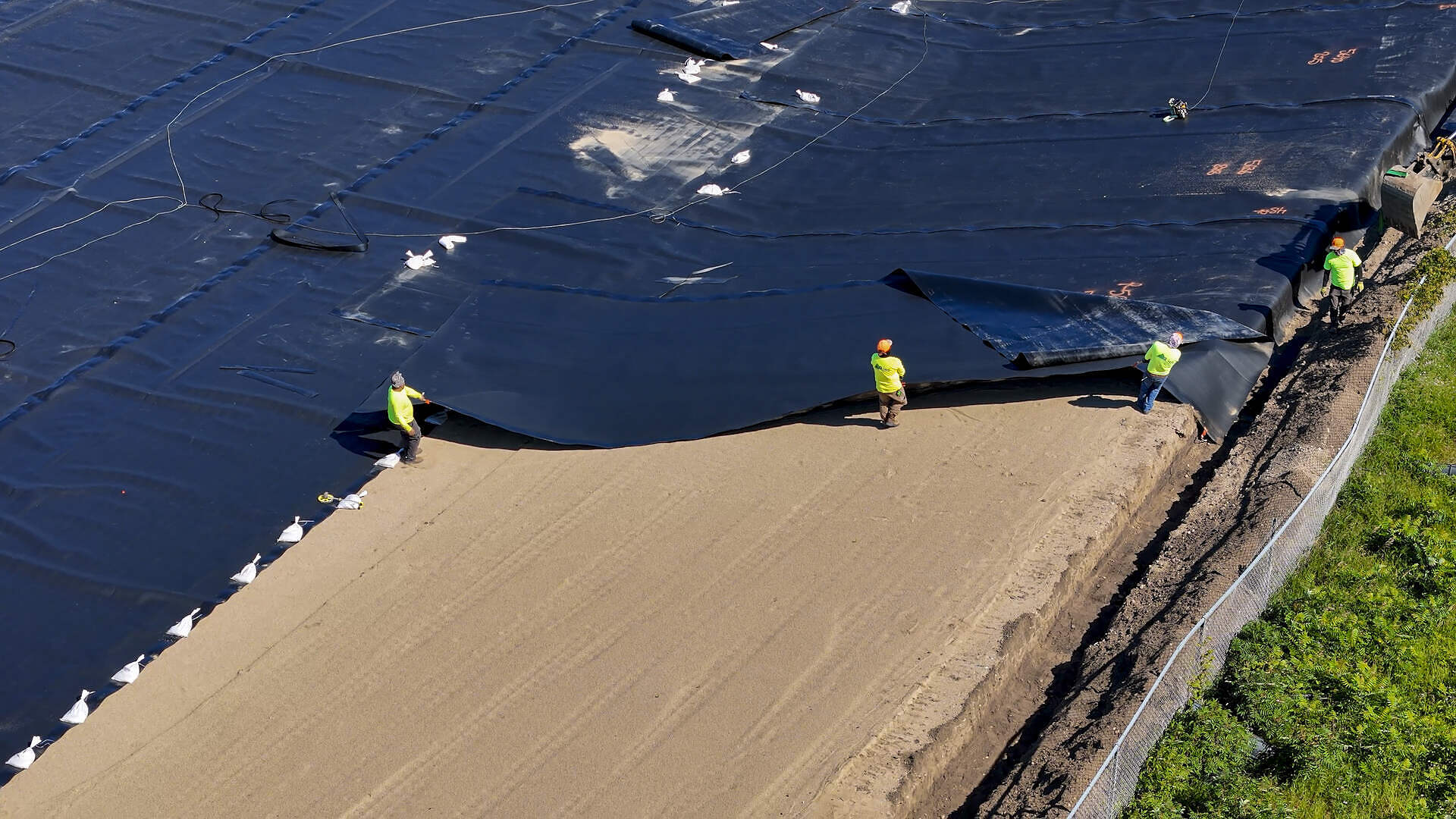 Three workers in safety gear install large black plastic sheeting over a sandy surface at a construction site, bordering an area with green vegetation, as part of preparations for constructing an Equalization Basin at the Water Resource Facility (WRF).