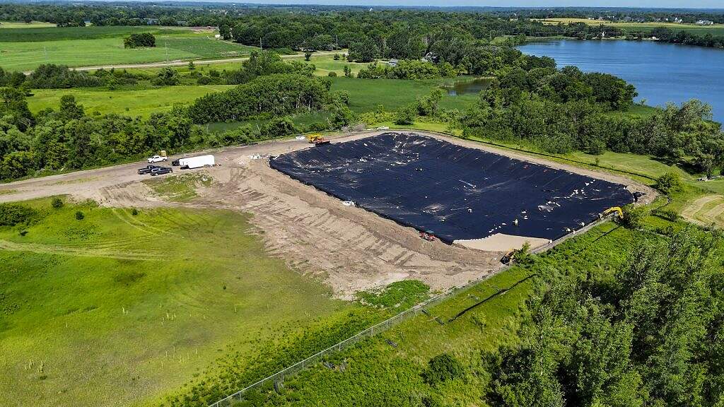 Aerial view of a large, black plastic-lined Equalization Basin at the WRF, surrounded by dirt and green fields, with construction vehicles and trees nearby and a lake visible in the background.