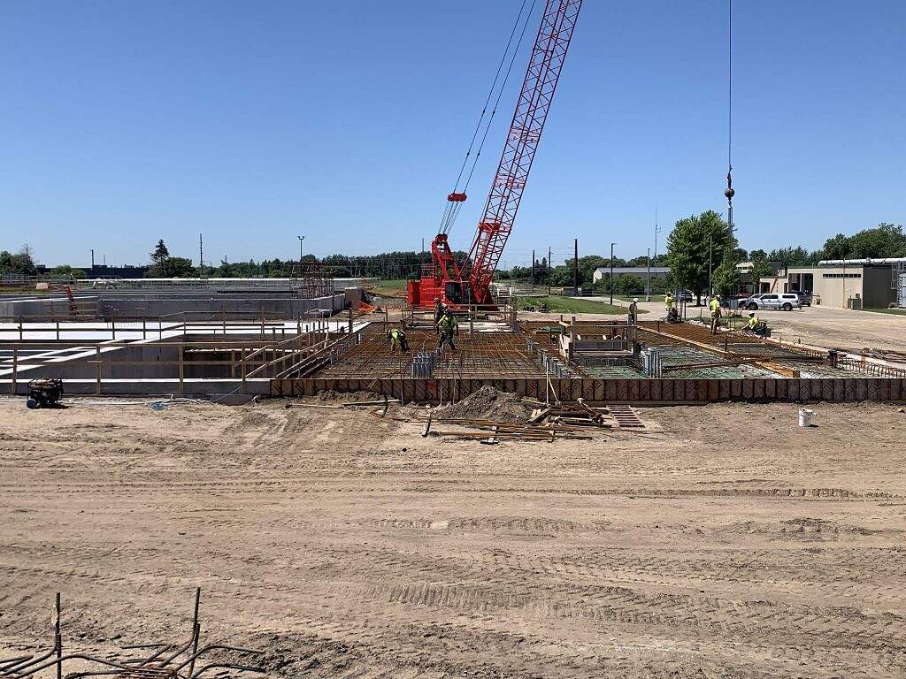 Construction site bustling with workers, WRF Improvements underway, dominated by a large red crane, steel reinforcements, and dirt ground beneath a clear blue sky. Some vehicles and buildings are visible in the background, highlighting ongoing construction activities.