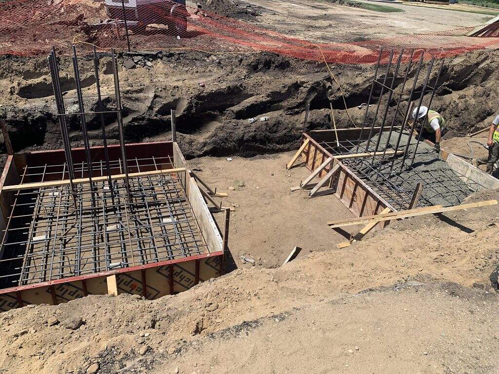 Two construction workers pour and smooth concrete into large rectangular footings with steel rebar, surrounded by dirt and excavation, at a construction site making WRF improvements. Orange safety fencing is visible in the background, emphasizing the focus on construction safety and infrastructure enhancement.