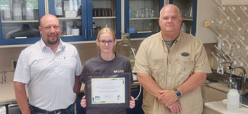 Three people stand in a laboratory. The person in the center holds a framed certificate for Wastewater Program Recognition. Shelves with lab equipment and bottles are visible in the background, highlighting their contributions to Minnesota Wastewater Monitoring.